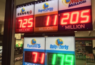 A signboard inside a convenience store displays the current jackpot amounts for various lottery games. There are bright, illuminated numbers in red and green showing the prize amounts: Powerball is at 725 million, Super Lotto Plus at 11 million, Mega Millions at 205 million, Fantasy 5 at 71 thousand, and Daily Derby at 179. In the background, shelves with various drinks and products are visible, along with promotional posters.